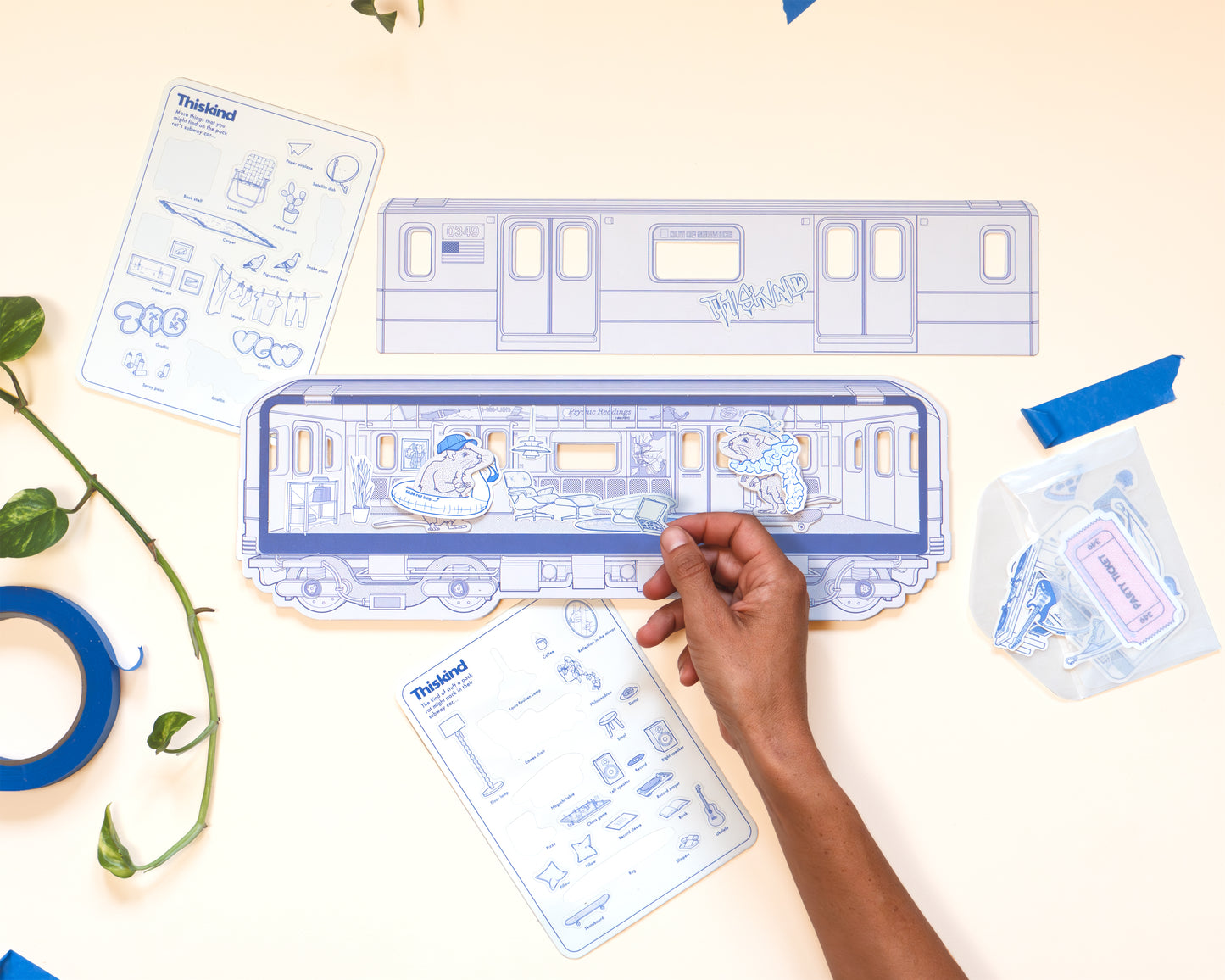 Paper craft detail image during play. Card stock subway car that opens to reveal subway interior with two subway rats dressed in paper hats and accessories. Shows the hand of the product user decorating the subway car with stickers that include chairs, lamps, plants pizza and more.